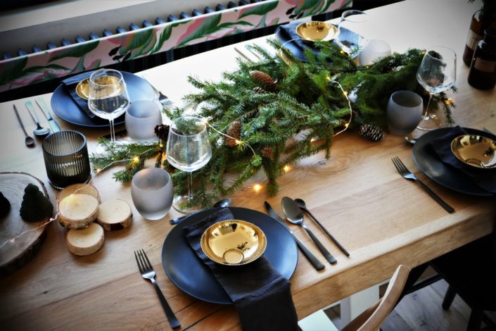 Table de Noël ambiance sous bois réalisée par Studio Candy pour For Me Lab - assiettes noir mat, bols dorés brillant, serviettes en lin, branches de sapin, boules de Noël en papier, pommes de pin