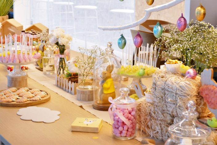 Goûter de Pâques naturel et pastel avec bottes de foin, oeufs en chocolat, bonbons, cake pops, fleurs, lapins et poules, nids meringue, sablés décorés