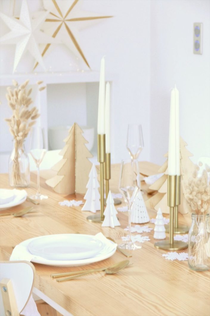 Table de Noël ambiance Laponie avec assiettes en céramique blanche, serviettes en lin blanc, flocons de neige, sapins en bois, bougeoirs dorés, fleurs séchées, couverts dorés mat