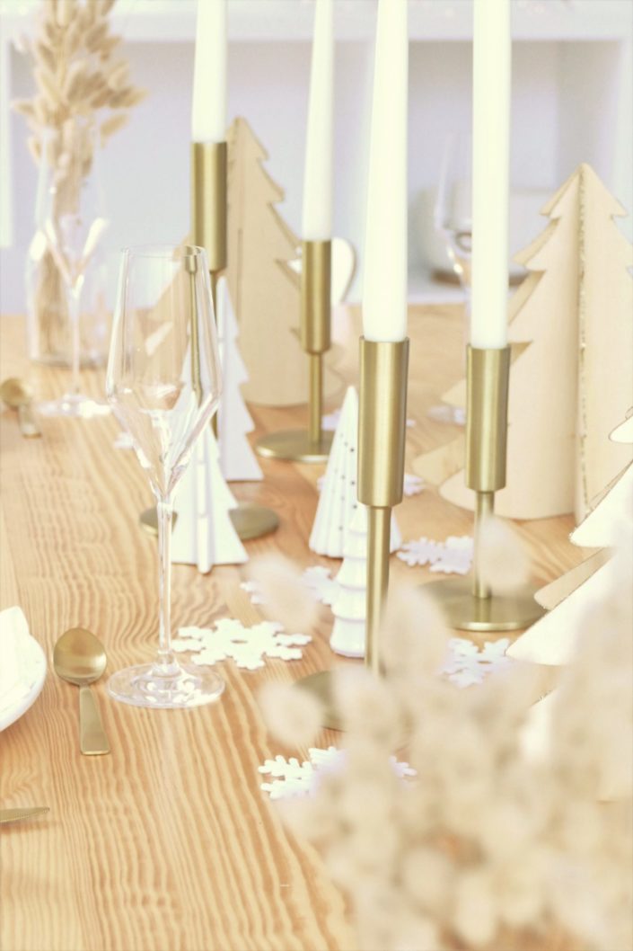 Table de Noël ambiance Laponie avec assiettes en céramique blanche, serviettes en lin blanc, flocons de neige, sapins en bois, bougeoirs dorés, fleurs séchées, couverts dorés mat