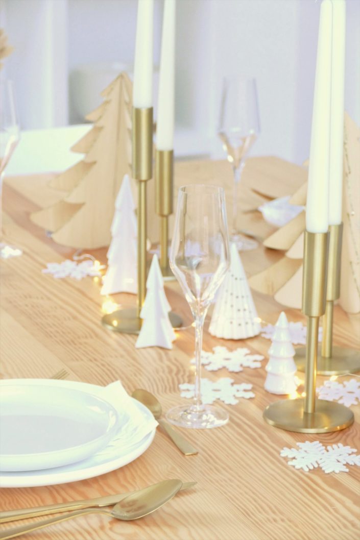 Table de Noël ambiance Laponie avec assiettes en céramique blanche, serviettes en lin blanc, flocons de neige, sapins en bois, bougeoirs dorés, fleurs séchées, couverts dorés mat