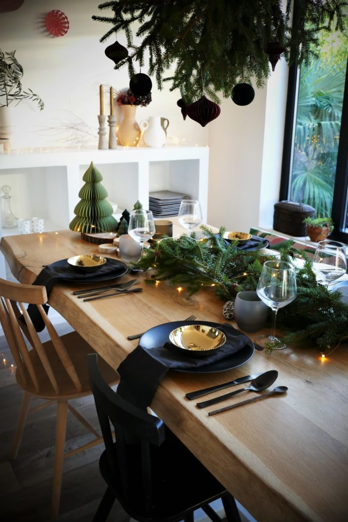 Table de Noël ambiance sous bois réalisée par Studio Candy pour For Me Lab - assiettes noir mat, bols dorés brillant, serviettes en lin, branches de sapin, boules de Noël en papier, pommes de pin