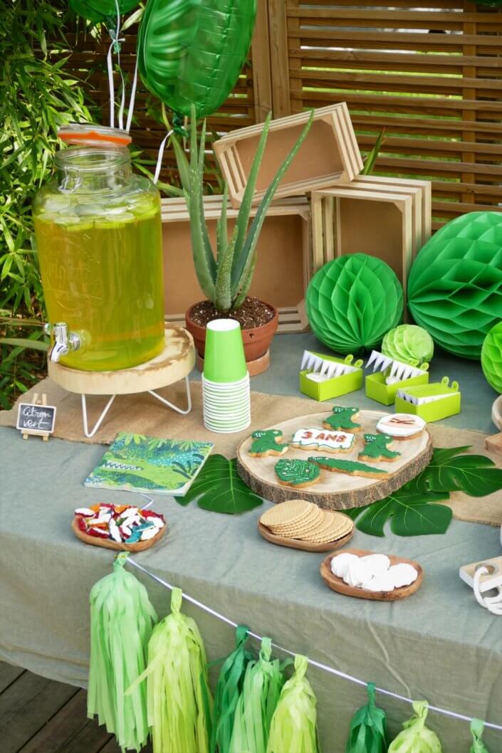 Anniversaire crocodile jungle avec sablés décorés , petits choux farcis, raisins, financiers amande, gâteaux au chocolat, bonbons. Décoration verte, feuilles tropicales monstera, boîtes crocodiles, vases, guirlande et ballons hélium.