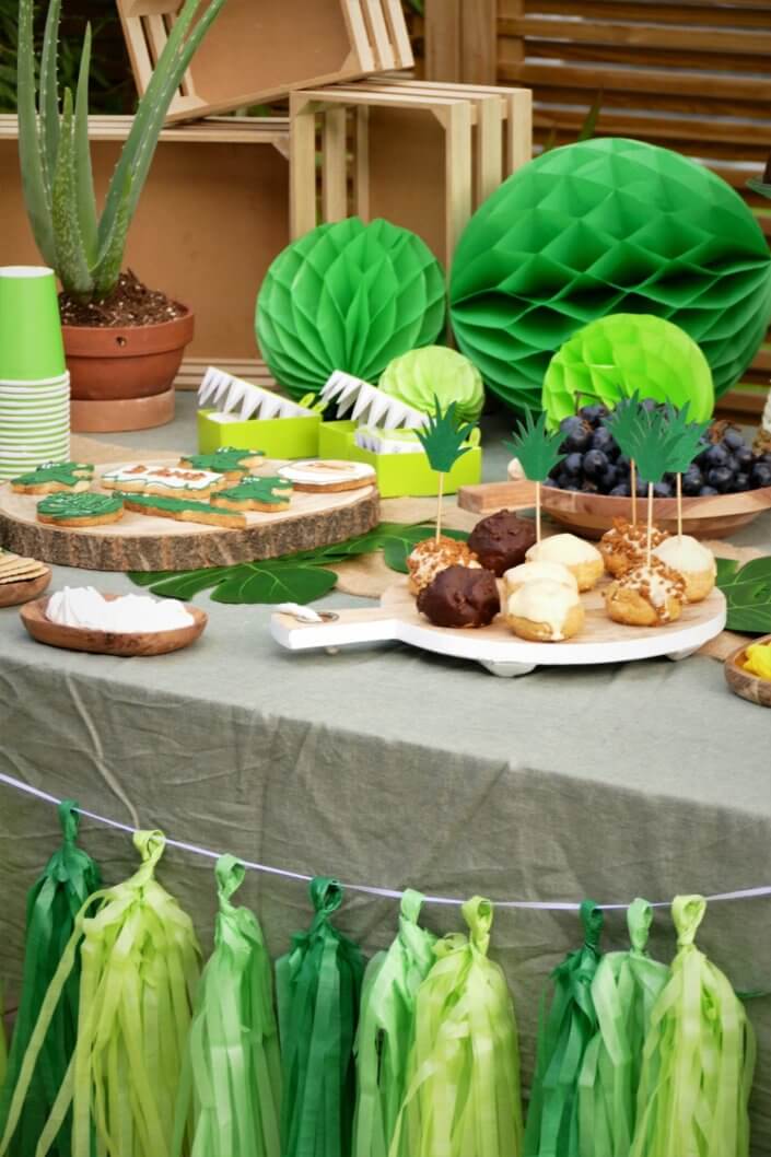 Anniversaire crocodile jungle avec sablés décorés , petits choux farcis, raisins, financiers amande, gâteaux au chocolat, bonbons. Décoration verte, feuilles tropicales monstera, boîtes crocodiles, vases, guirlande et ballons hélium.