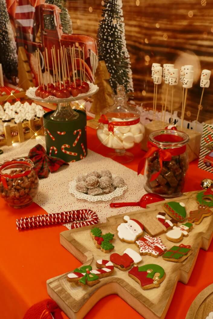 Goûter de Noël traditionnel avec petits sablés, cakepops rouges pailletés, chamallows bonhommes de neige, sapins meringue, petits oursons guimauve. Décoration en rouge, or, vert sapin avec des sucres d'orges, des bonbons, des sapins en bois