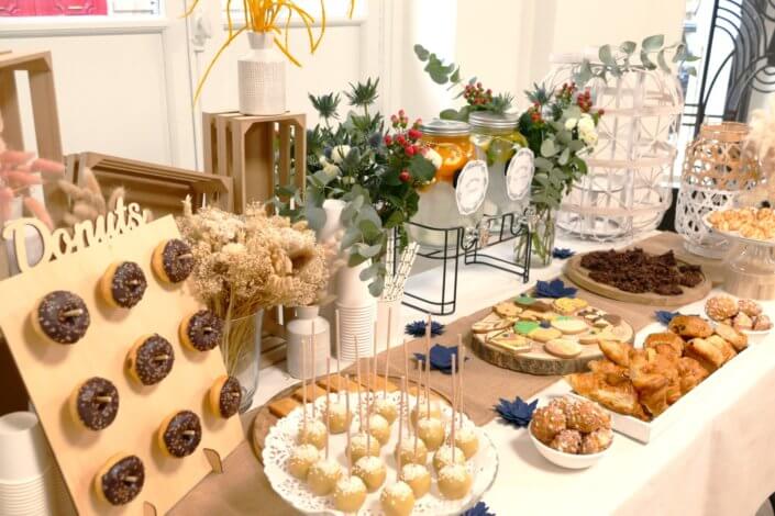 Réalisation d'une table gourmande décorée avec cake pops, sablés décorés, roses des sables, financiers, fleurs fraîches, fontaines à boissons d'eaux infusées, chemin de table en toile de jute, lanternes en bois.