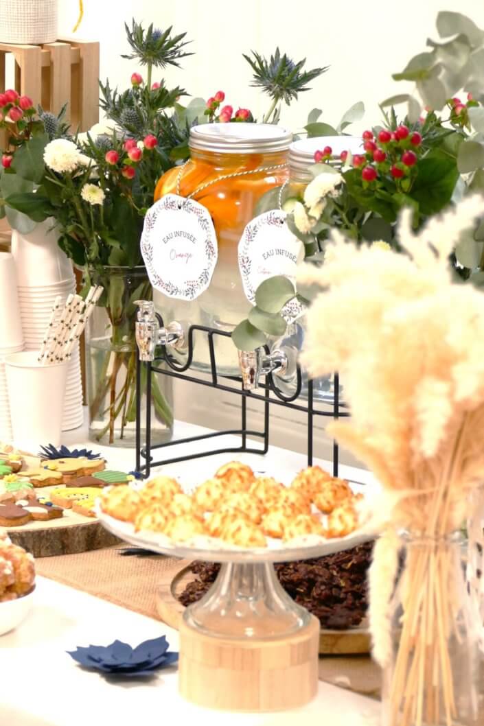 Réalisation d'une table gourmande décorée avec cake pops, sablés décorés, roses des sables, financiers, fleurs fraîches, fontaines à boissons d'eaux infusées, chemin de table en toile de jute, lanternes en bois.