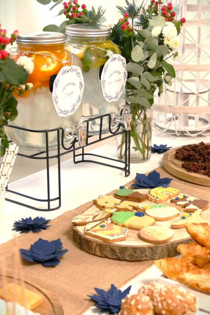 Réalisation d'une table gourmande décorée avec cake pops, sablés décorés, roses des sables, financiers, fleurs fraîches, fontaines à boissons d'eaux infusées, chemin de table en toile de jute, lanternes en bois.