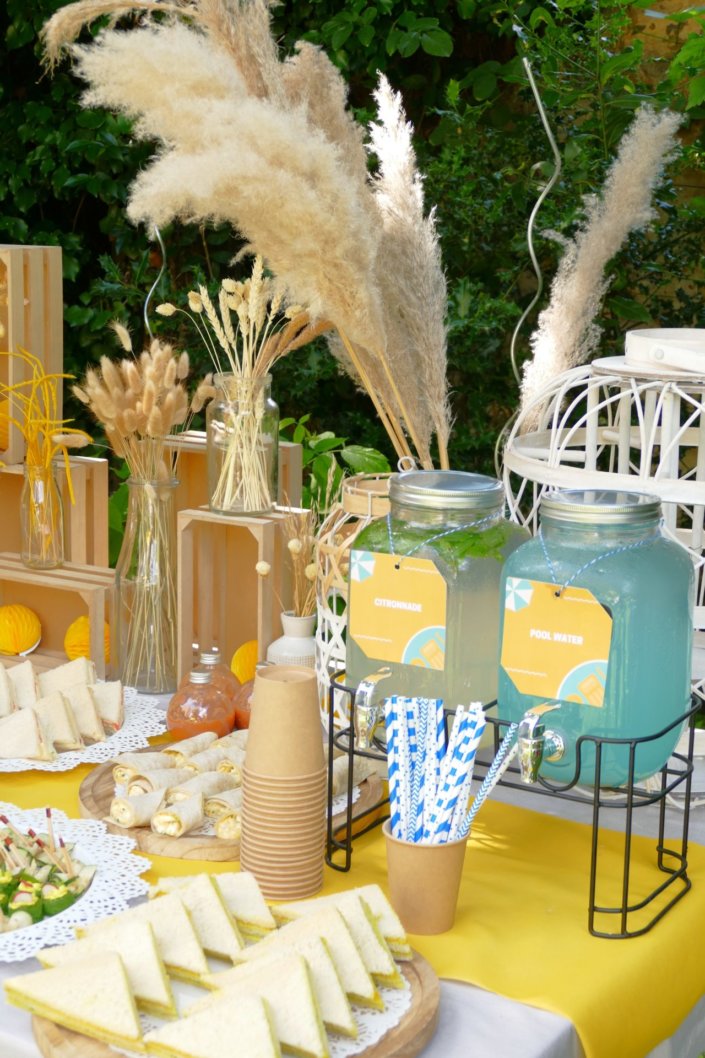 Pool party Uriage avec buffet décoré sur mesure de fleurs séchées, lanternes en bois, patisseries et sablés décorés en forme de crème solaire, cakepops, financiers, gâteaux