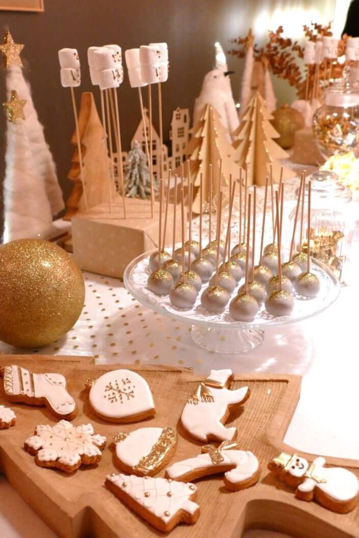 Soirée de Noël Adopt par Studio Candy avec une grande table de Noël décorée en or et blanc. Cakepops au chocolat, cupcakes, sapins meringue, sablés de Noël et corbeille de fruits et cocktail salé avec verrines, grazing platters, cônes garnis