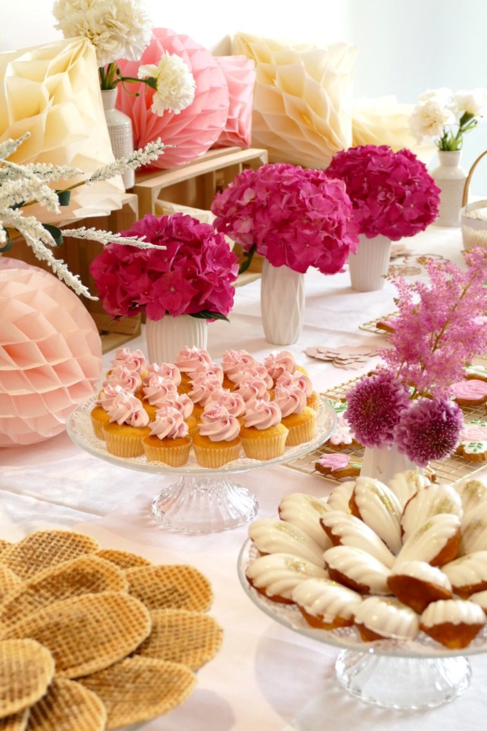 Goûter fleuri en rose et blanc réalisé sur mesure par studio candy avec des pâtisseries personnalisées, mini cupcakes, madeleines à coque rose, gaufres, sablés décorés, et scénographie printanière